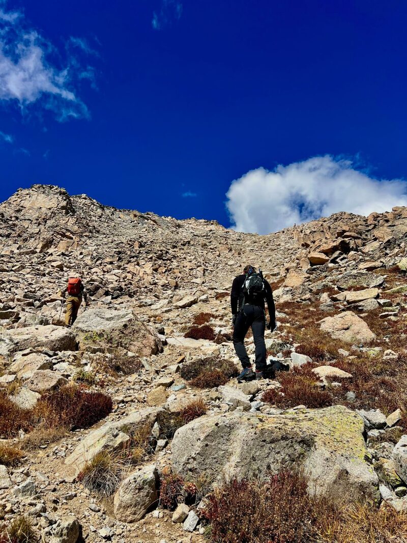 Two hikers climb a steep, rocky slope toward a ridge