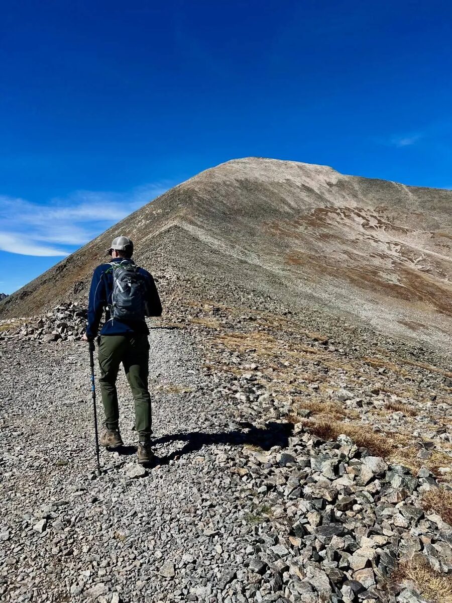 A hiker with a trekking pole walks along a rocky ridge toward a bare mountain summit