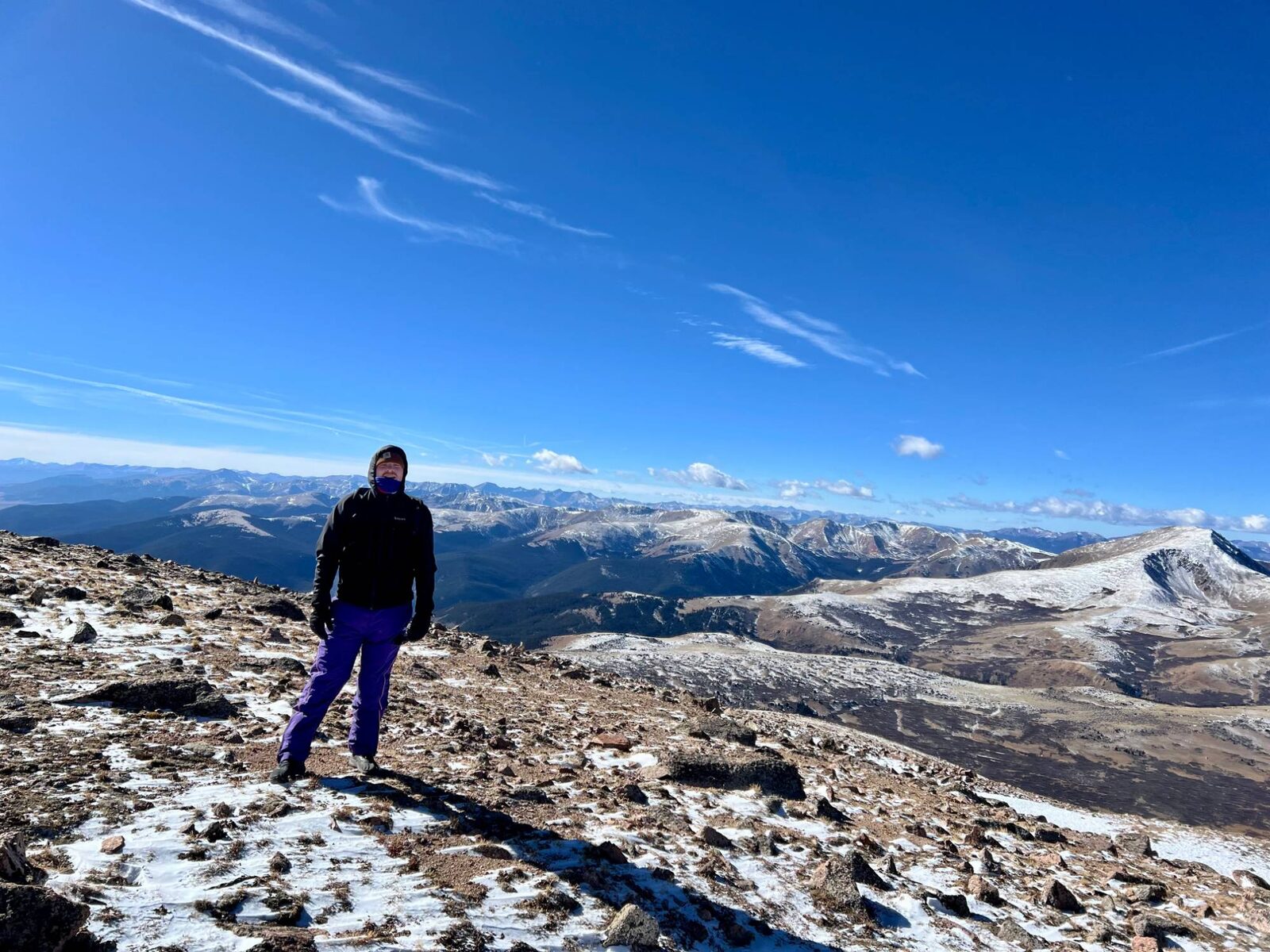 A man stands on a snowy, rocky summit above a wide mountain valley