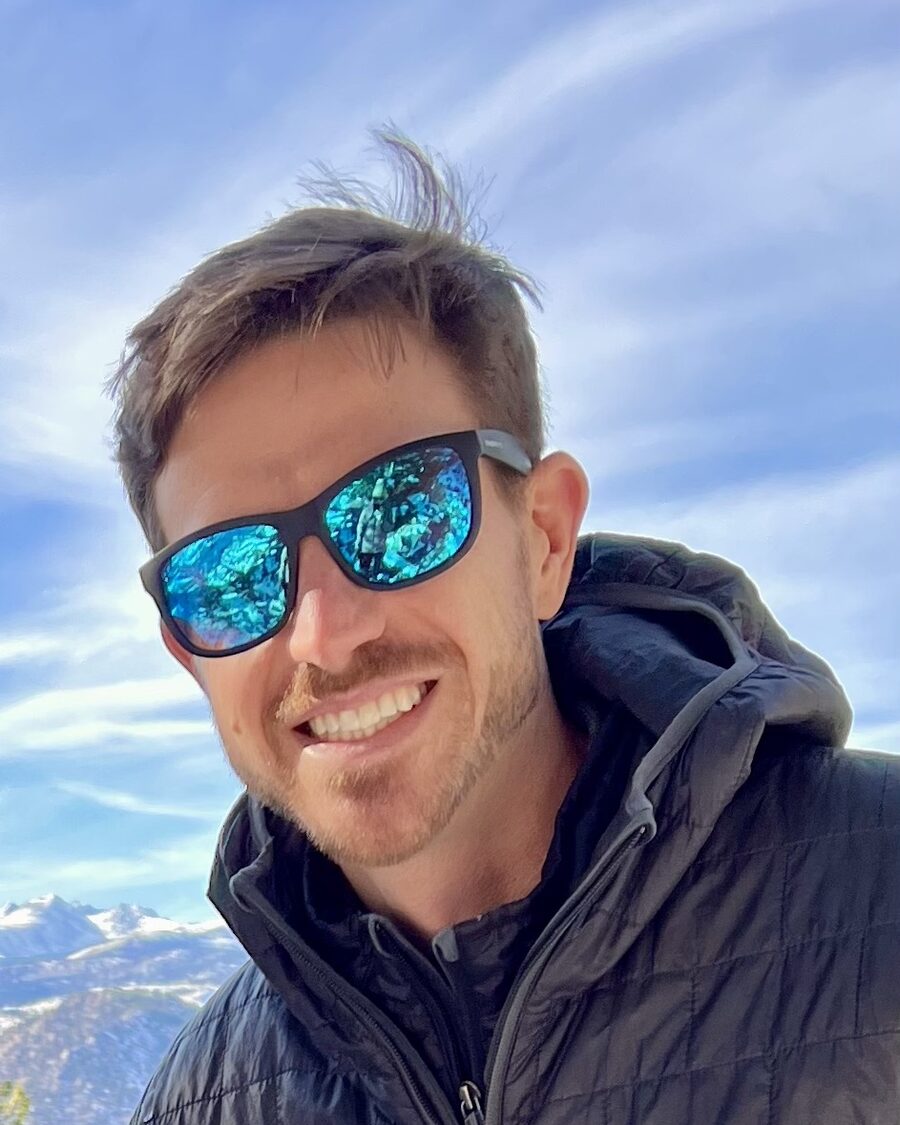 Travis smiling in sunglasses, with snow-capped mountains behind him